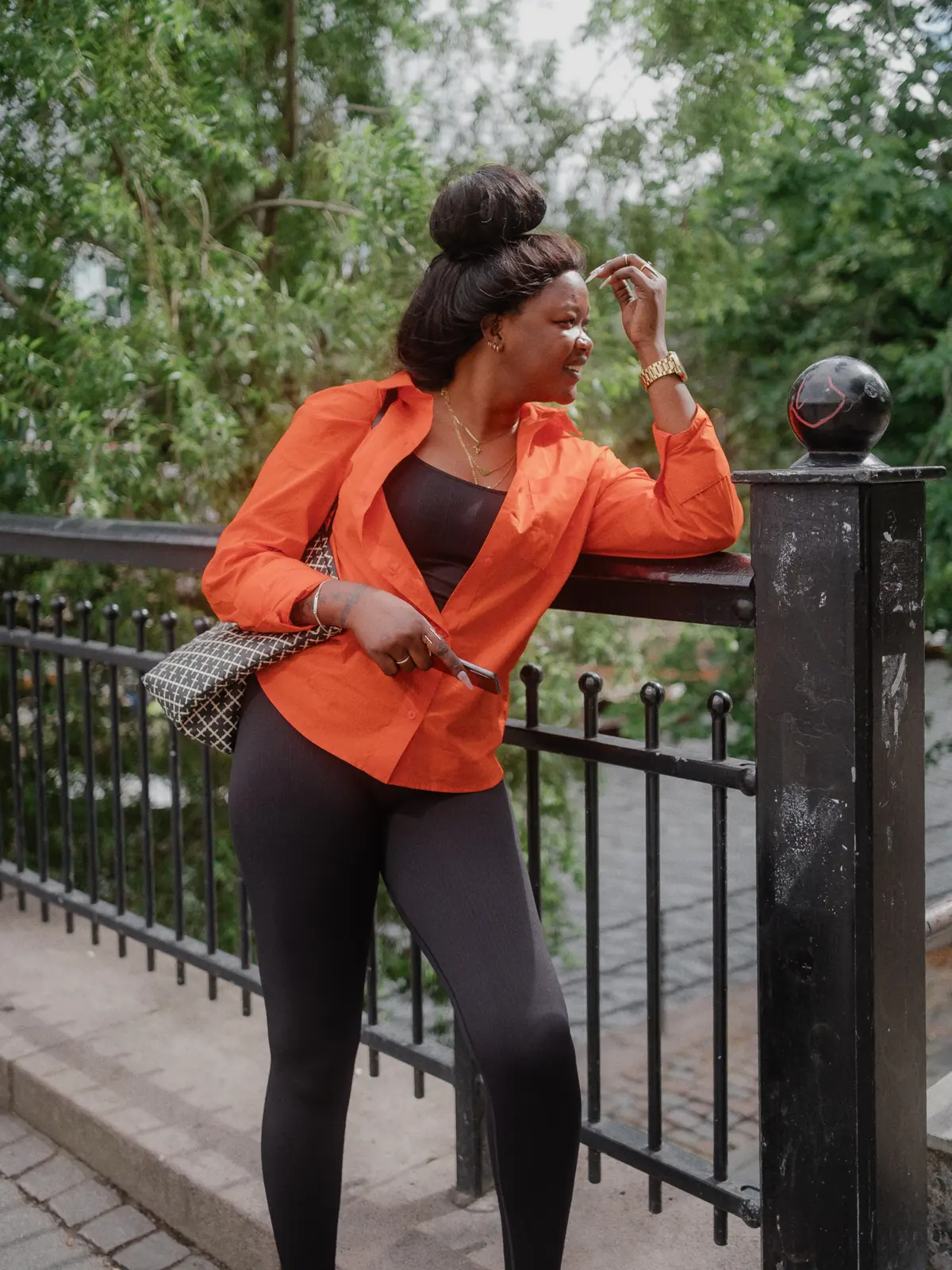 Street portrait of a black woman naturally posed against a bridge fence wearing an orange top agains a background of foliage.