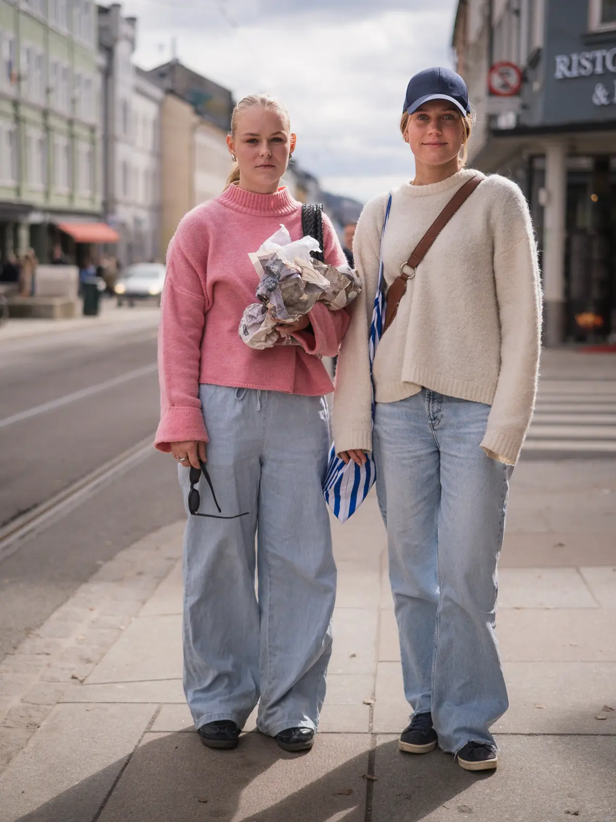 Two young women photographed on the street, both wearing light blue, baggy denim pants and sweaters in muted colours.