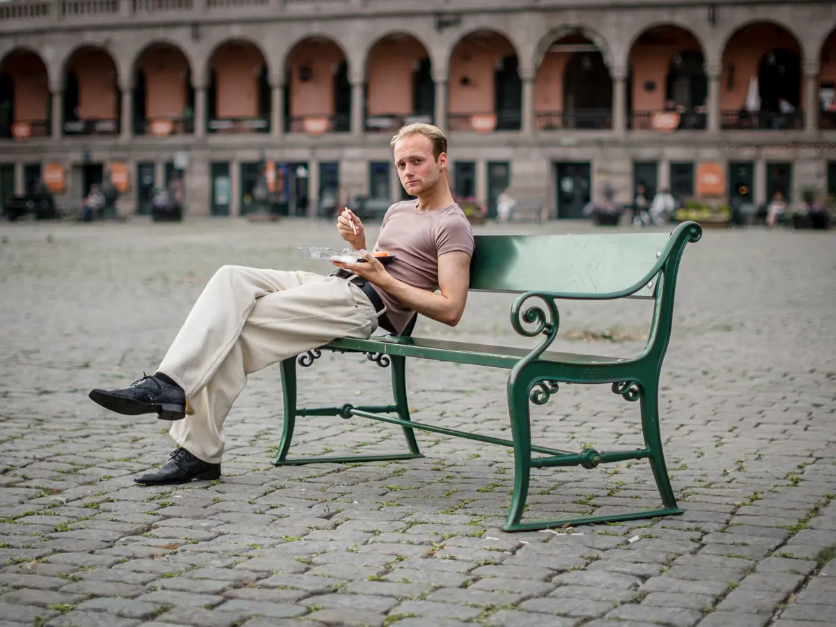Young man wearing muted colors having sushi on a green bench at Youngstorget.