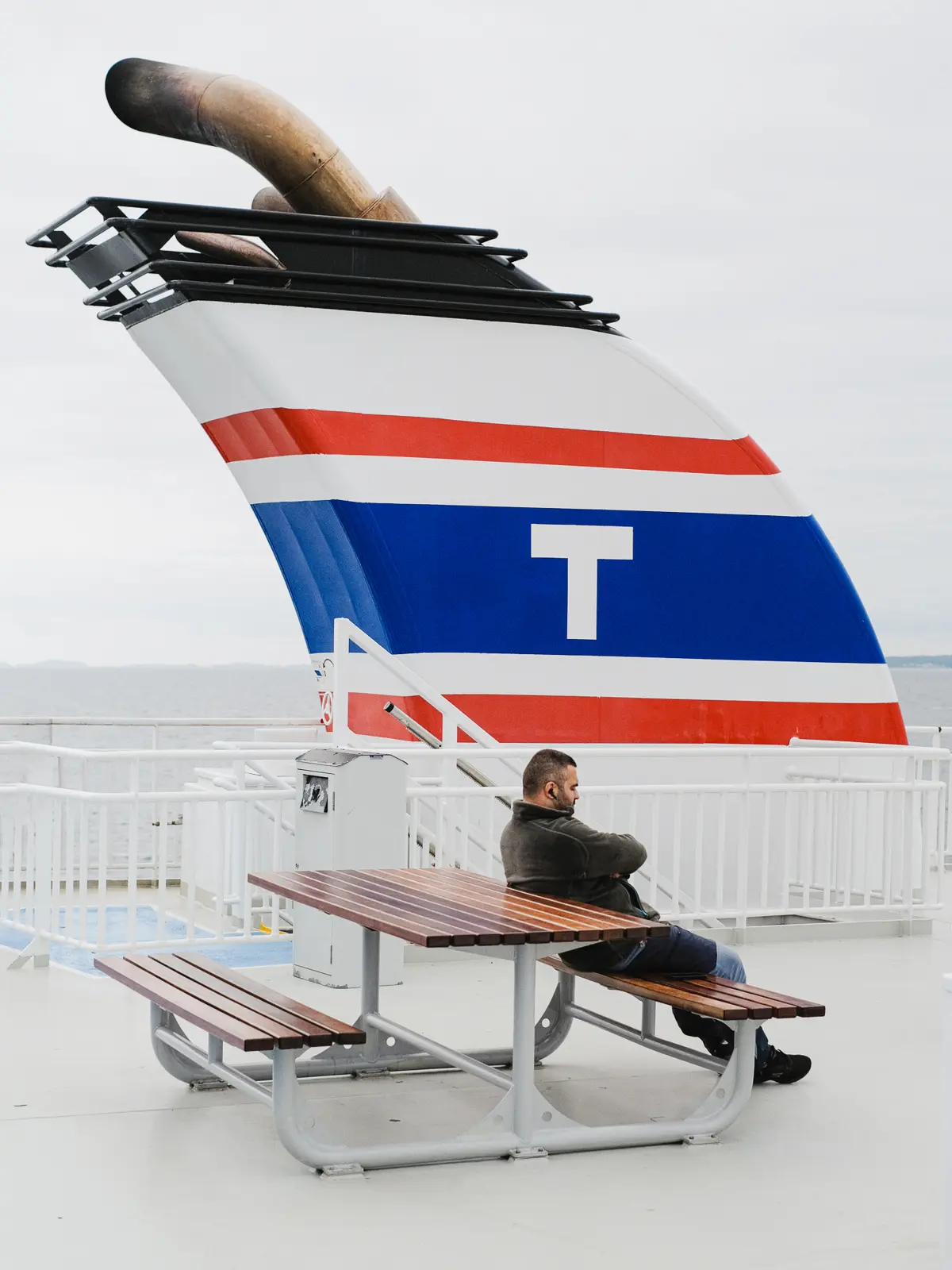 Man leaning back on a bench on a ferry with a large chimney in the background shaped opposite to his posture.