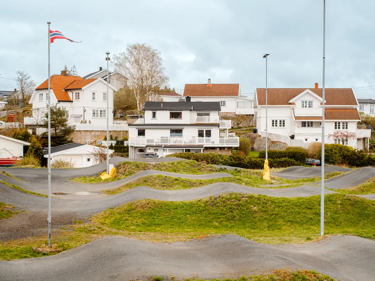 Wavy asphalt interspersed with grass and flag poles in the foreground. White houses in the background.