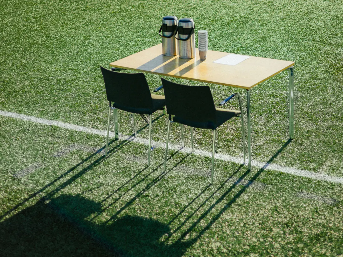 Small table with two chairs and two thermoses of coffee placed on the white line of a soccer pitch.