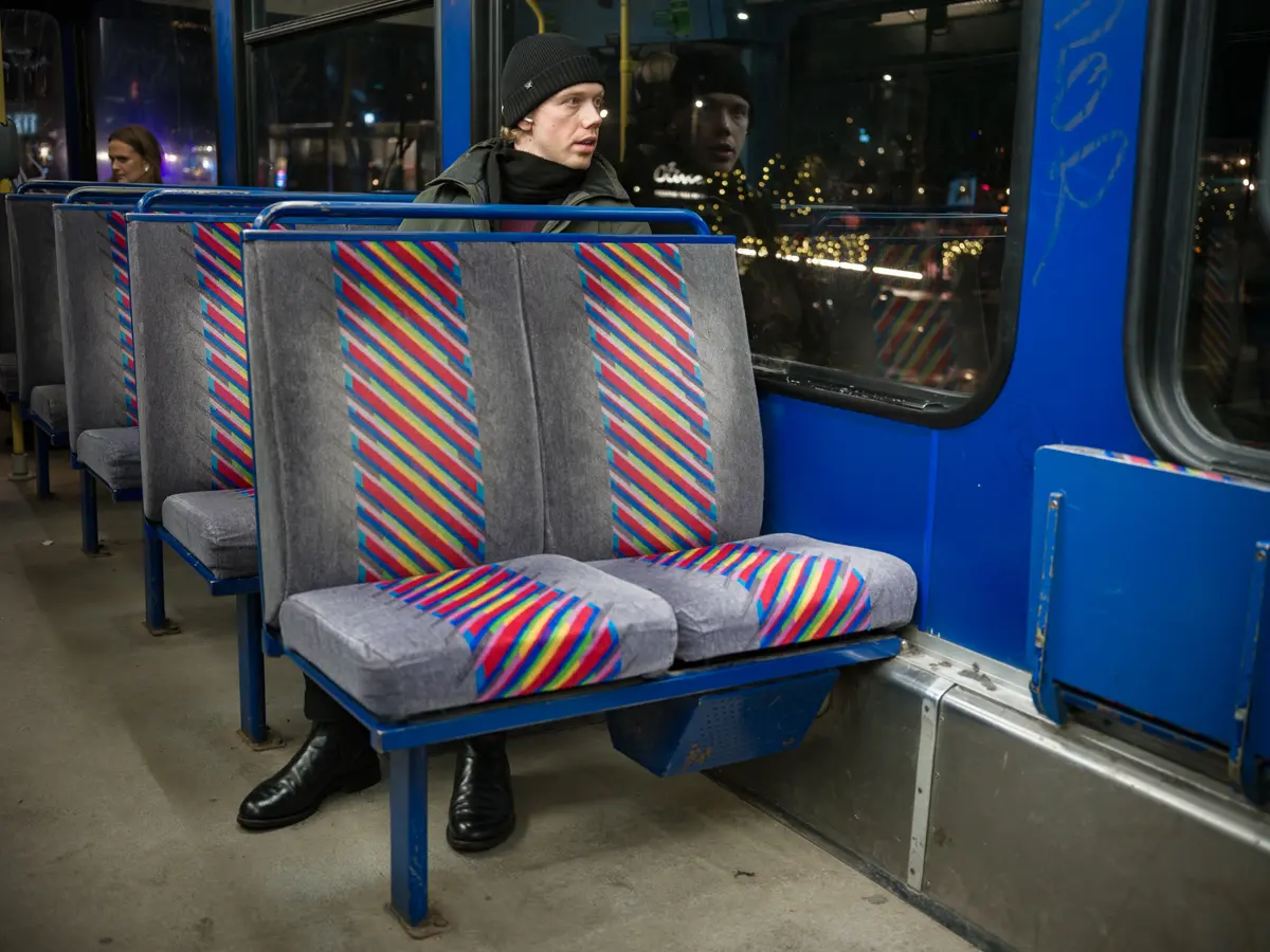 Young man seated alone on the back left isle of seats, on the second row. Only his head and feet are visible. His feet sticking out under the seats. Seems like the tram is designed for shorter people than him.