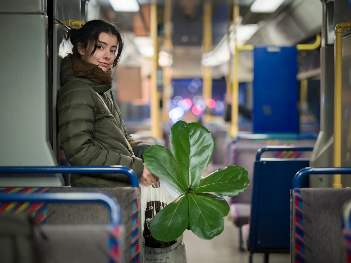 Young woman standing on the tram, her body facing right but her head facing the camera. She carries a large, green plant.