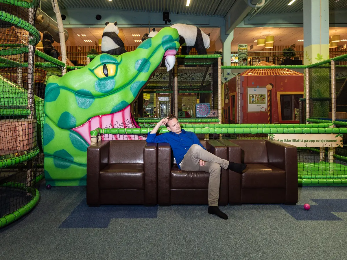 Man sitting in brown leather sofa attending children at an indoor playground. Behind him is an enormous crocodile head with its mouth gaping over him.