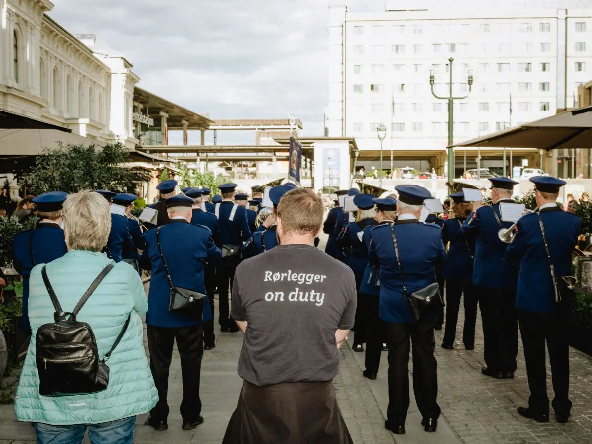 Crowd with marching band photographed from behind. Foreground shows a man's back with a T-shirt that says "Plumber on duty" in Norwegian.