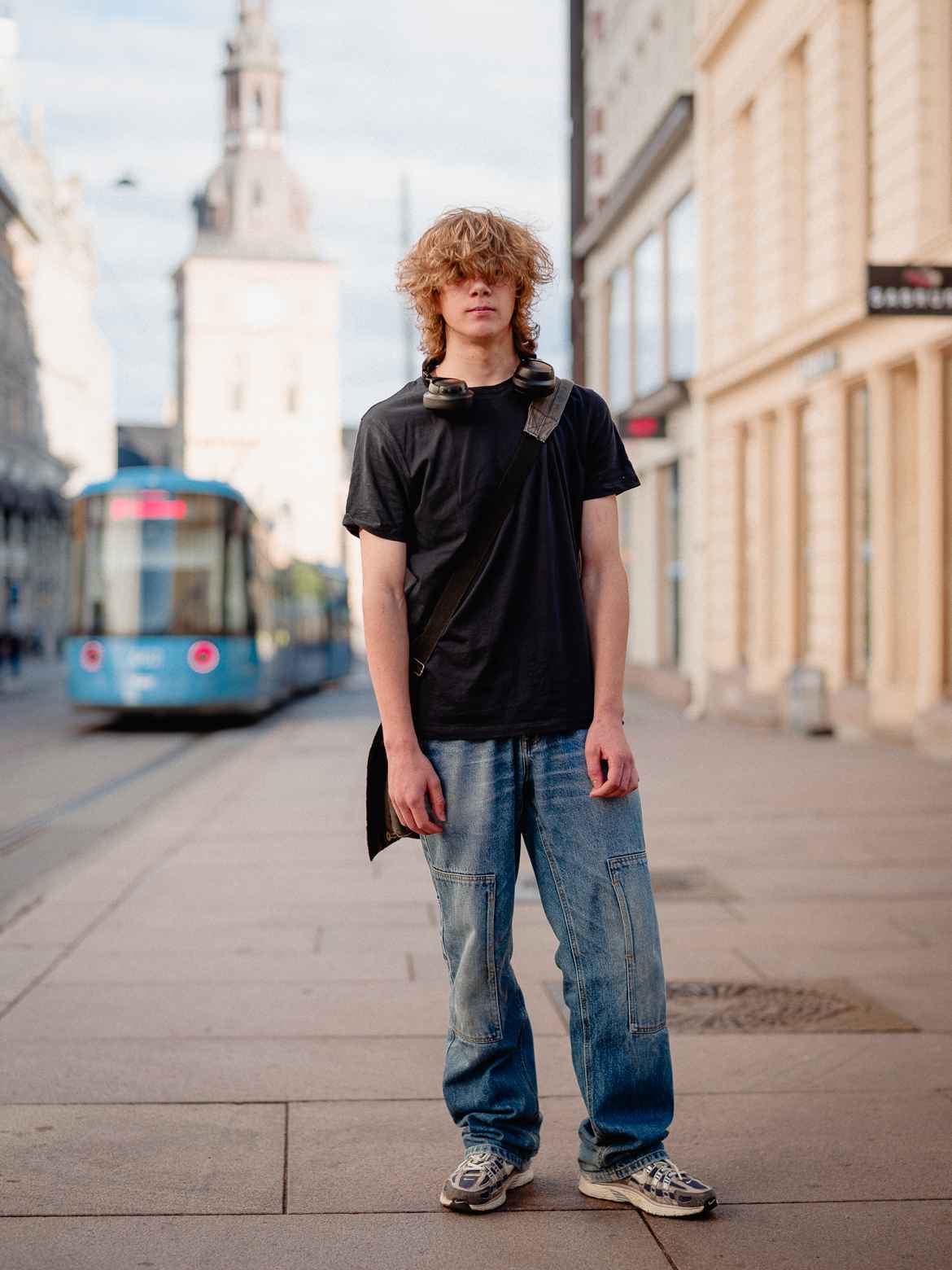 Young man in baggy jeans and black T-shirt with long, curly bangs covering his eyes photographed on the street in Oslo