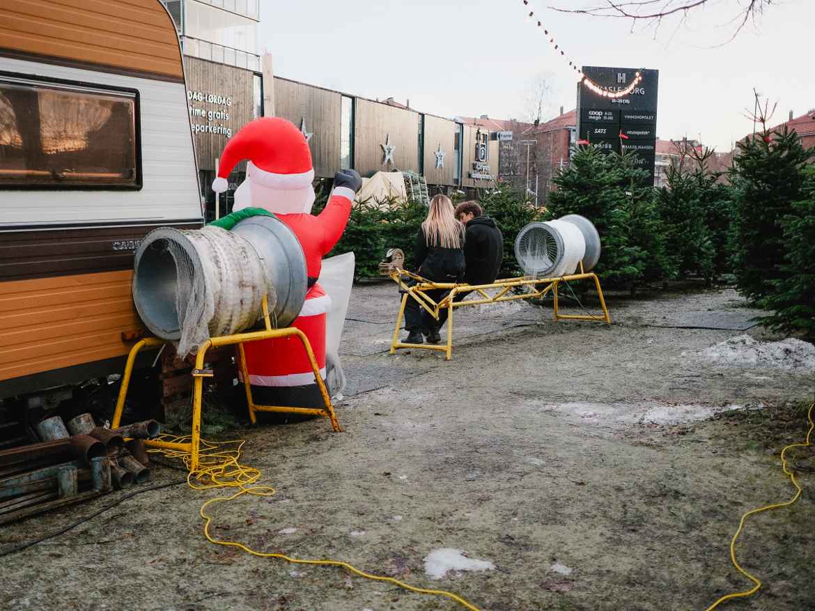Temporary outdoor sales lot for Christmas trees decorated with an inflatable santa against the packing devices, yellow cable on the ground circling the lot and a young couple waiting in line.