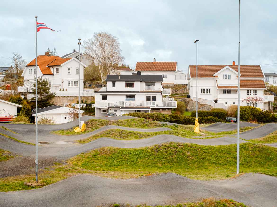 Wavy asphalt interspersed with grass and flag poles in the foreground. White houses in the background.