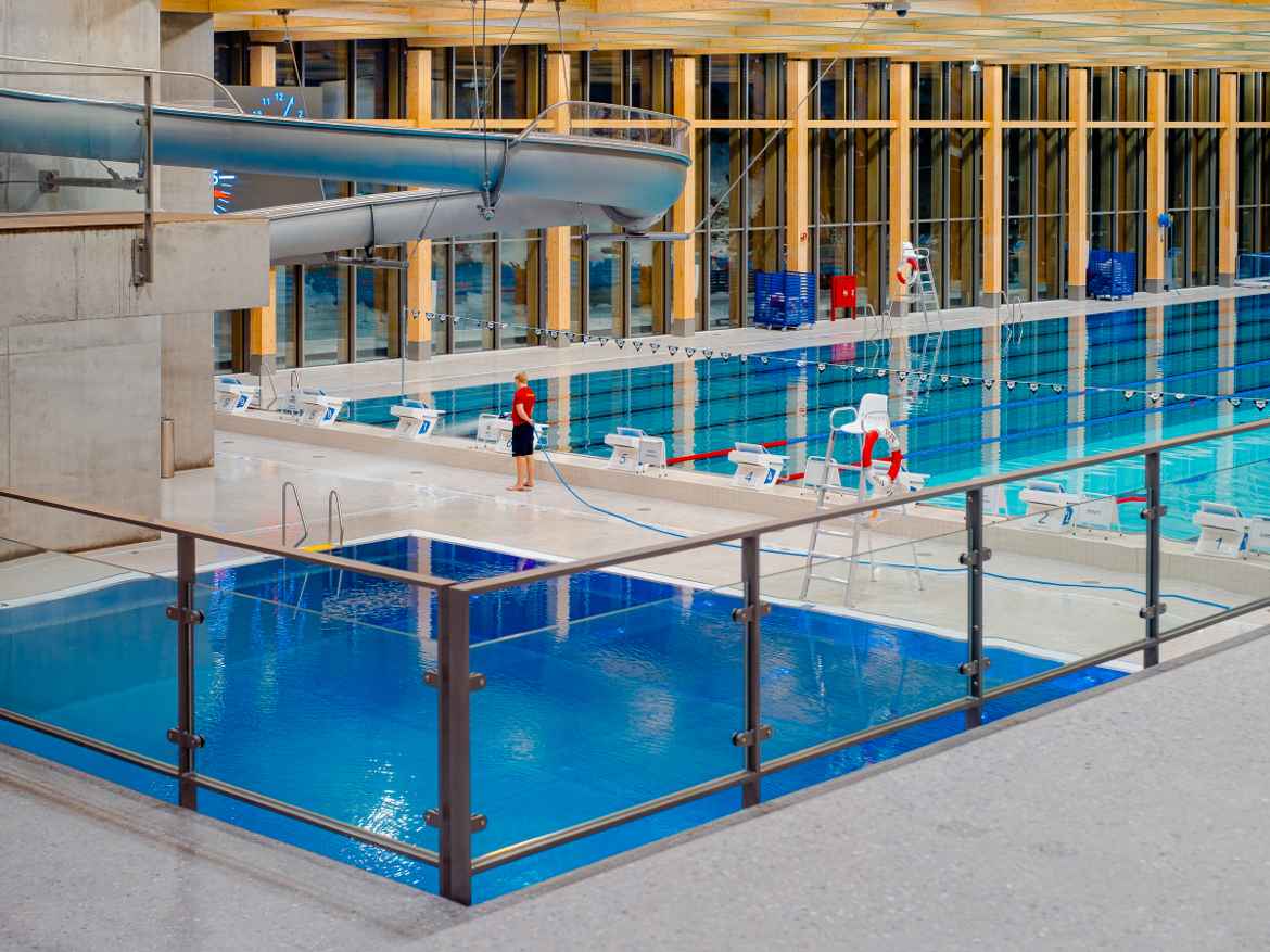 Tøyen indoor swimming complex after closing time. The water is blue an calm and a lone man drags a long, blue hose across the floor.