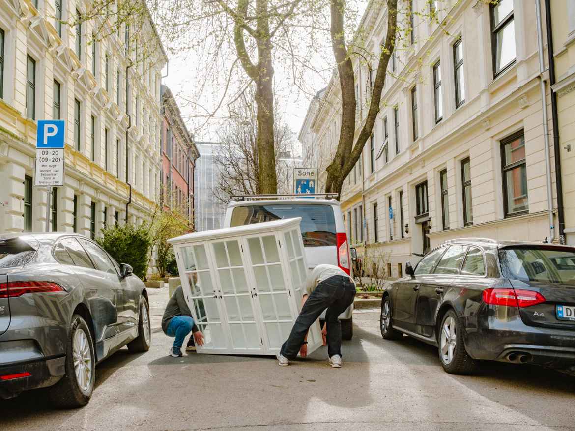Two men trying to lift a glass display cabinet in the street between four story apartment buildings that look similar to the cabinet