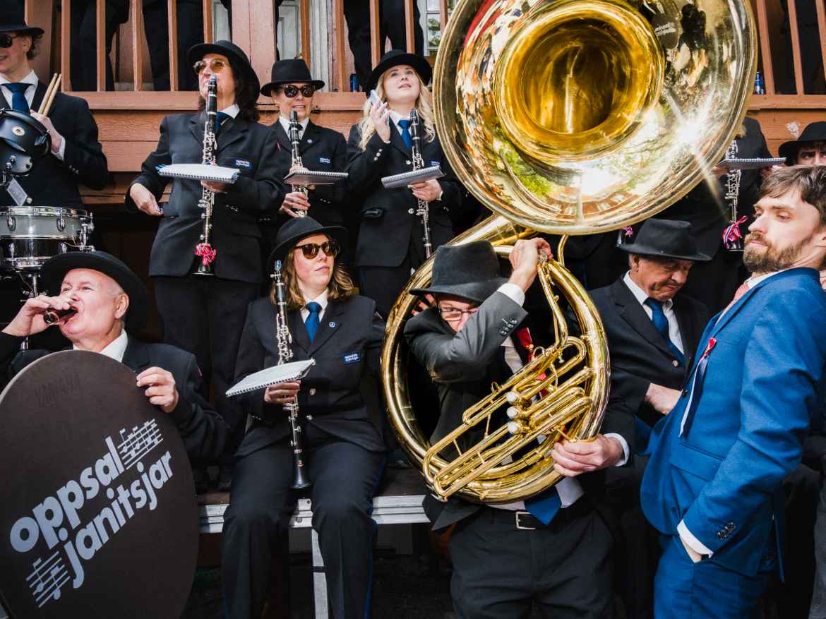 Oppsal Janitsjar marching band playing at Asylet, Oslo on constitytion day in a chaotic scene where one man is drinking from a shot glass wile another tries donning a huge instrument almost knocking over a man trying to pass him.