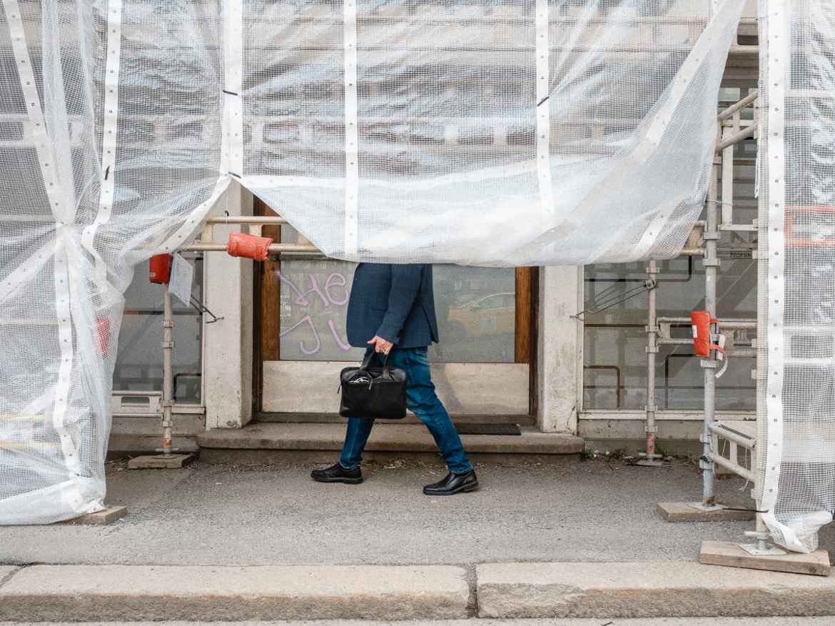Man in blue blazer and jeans carrying a black briefcase behind scaffolding with construction draping covering his head