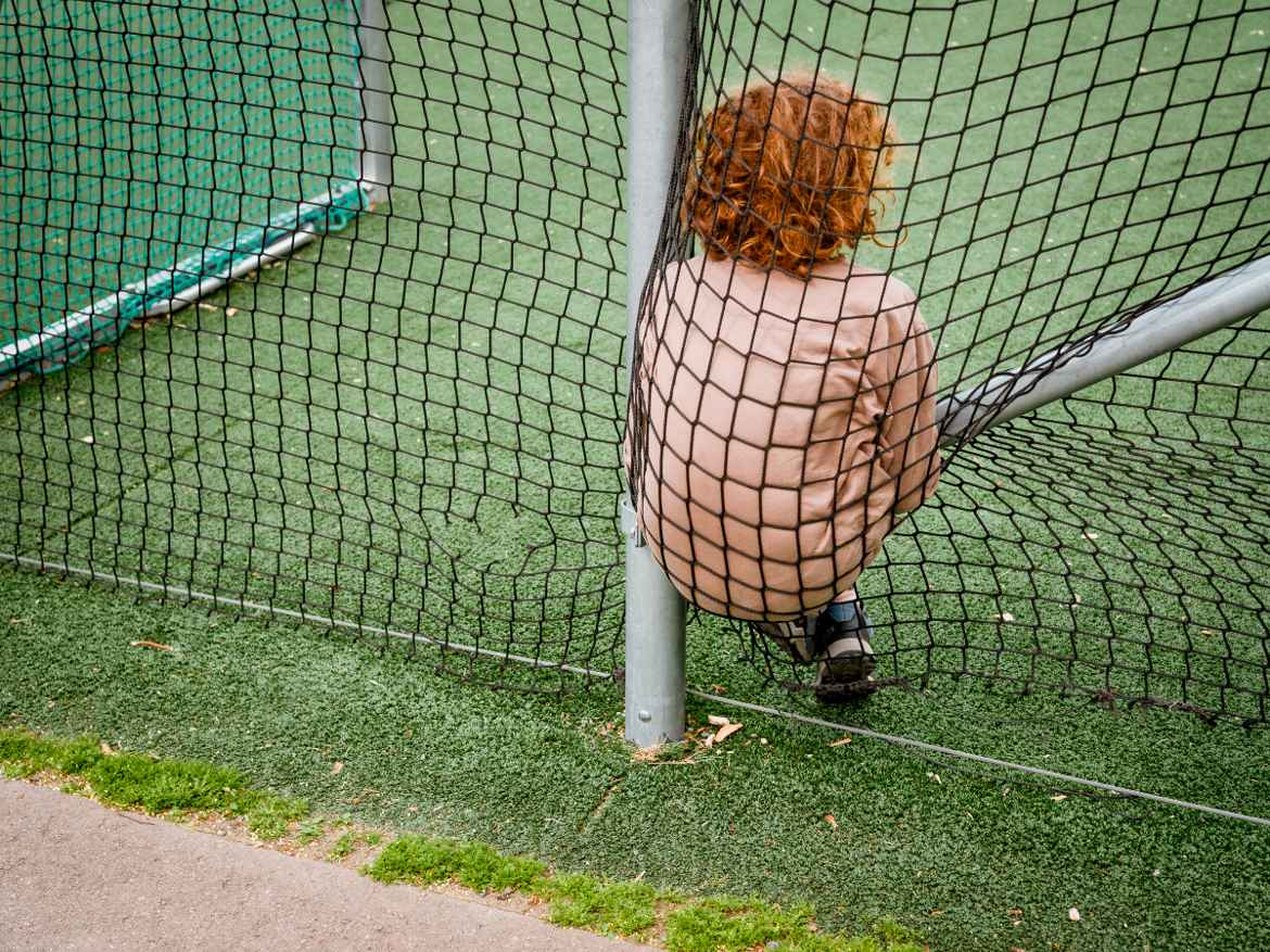 Ginger boy shot from behind in orange jacket using the net around a neighbourhood football pitch as a chair