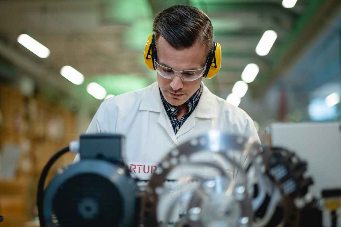 Young man in white lab coat with eye and ear protection looking down and operating heavy machinery.