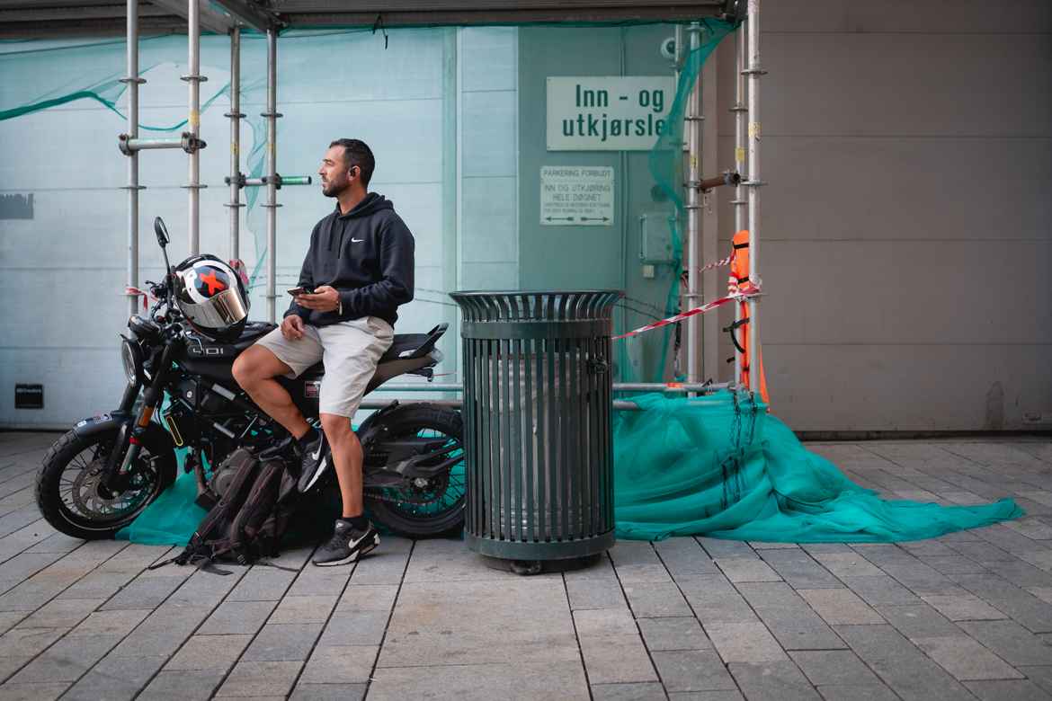 Man sitting sideways on the seat of his parked motorcycle next to a street trash can with construction draping flowing around him on the ground.