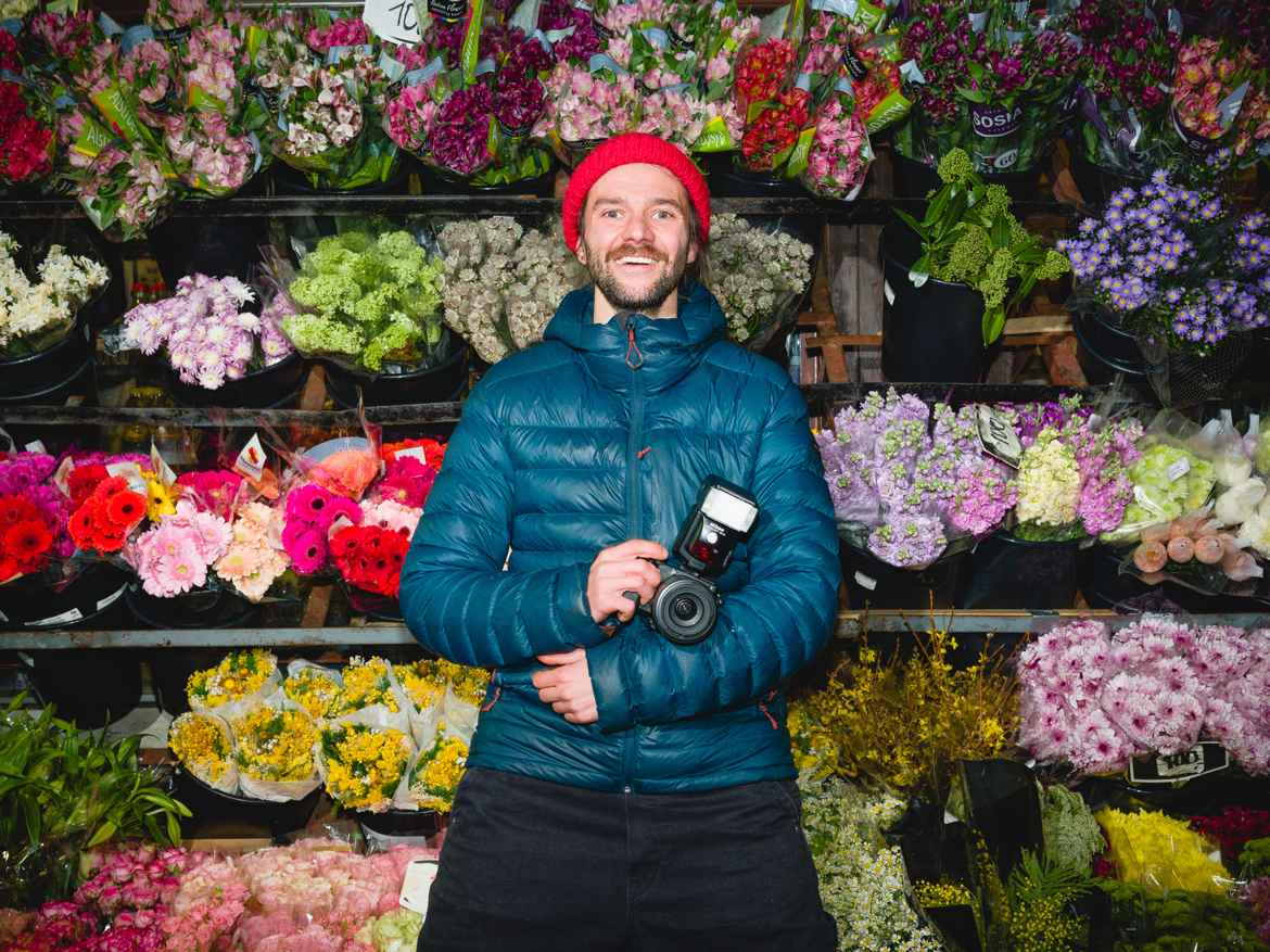 Photographer Dawid Łozowicki photographed smiling with flash against a wall of colorful flowers.