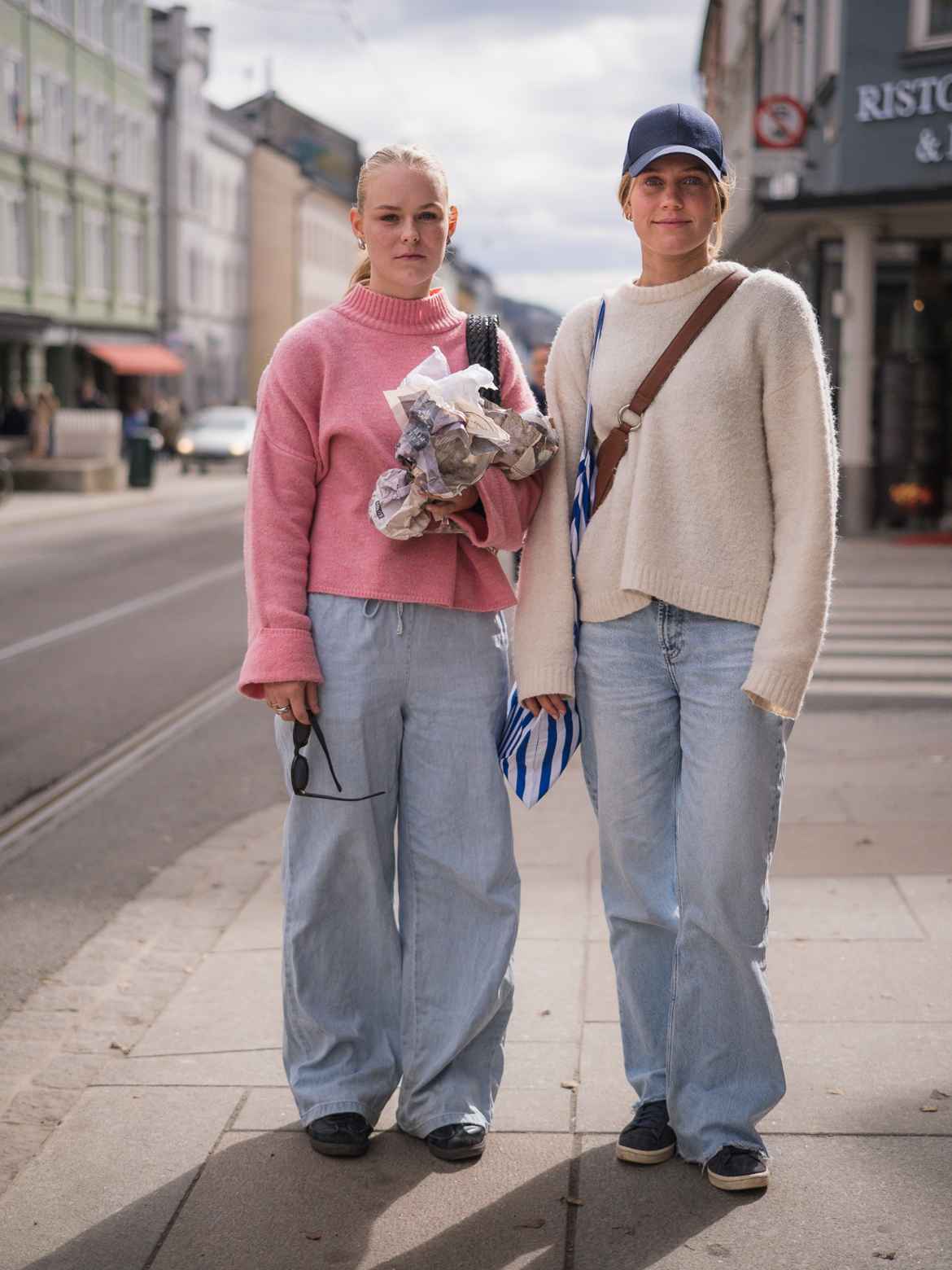 Two young women photographed on the street, both wearing light blue, baggy denim pants and sweaters in muted colours.