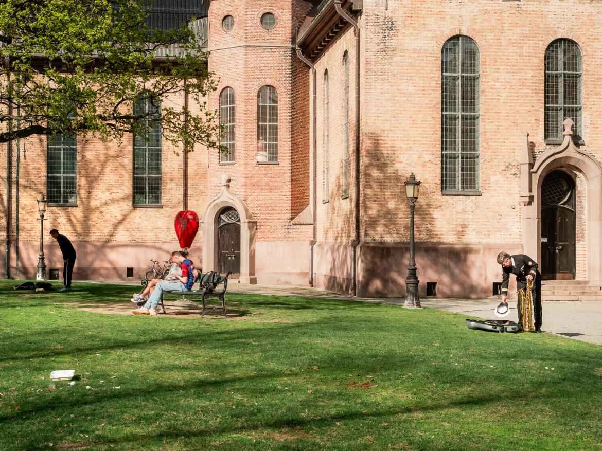 A sunny day in May outside Oslo Domkirke where a teen couple sits together on a bench under a huge, red heart while a man in marching band uniform looks to be packing up his bass saxophone on the lawn next to them.