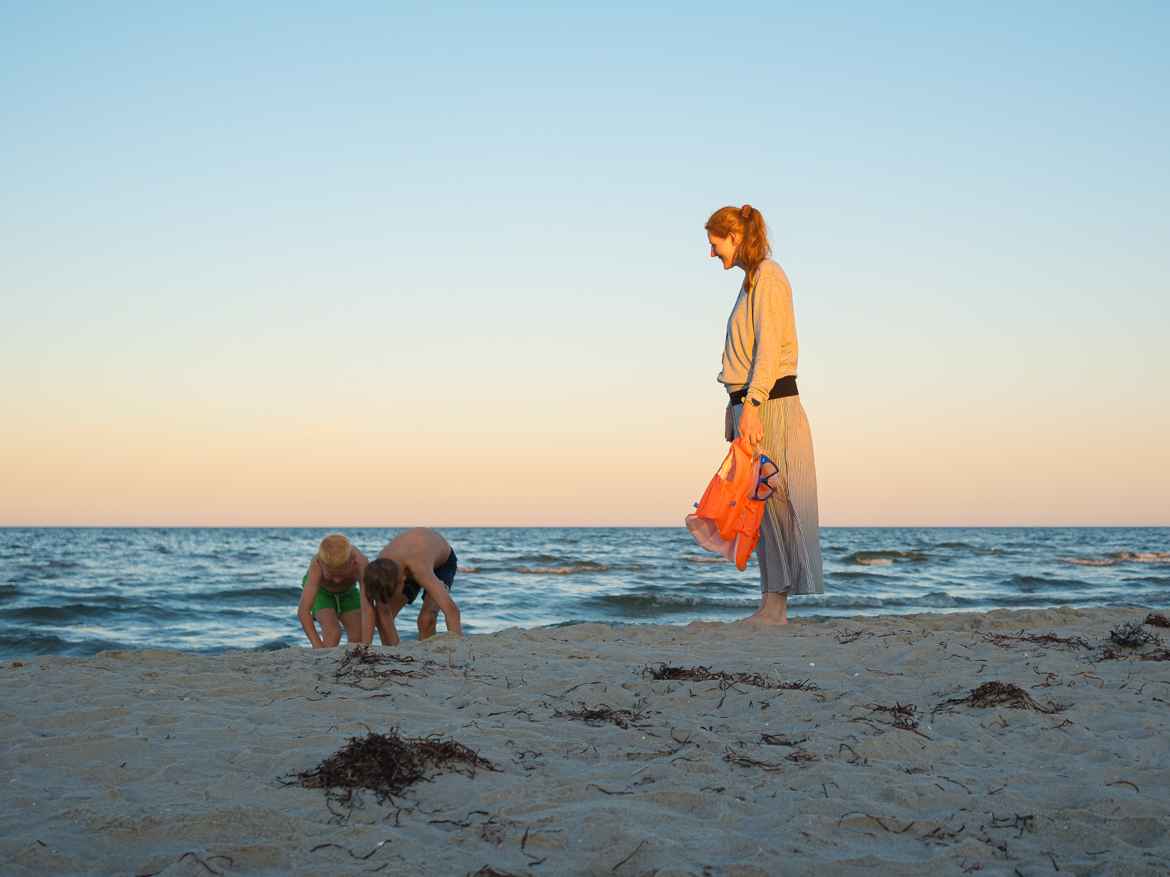 Two boys playing in the sand on a beach while their mother stands above them patiently waiting for them to join her home. The sun is going down which is evident by the shadow from the horizon hitting the woman's lower half.