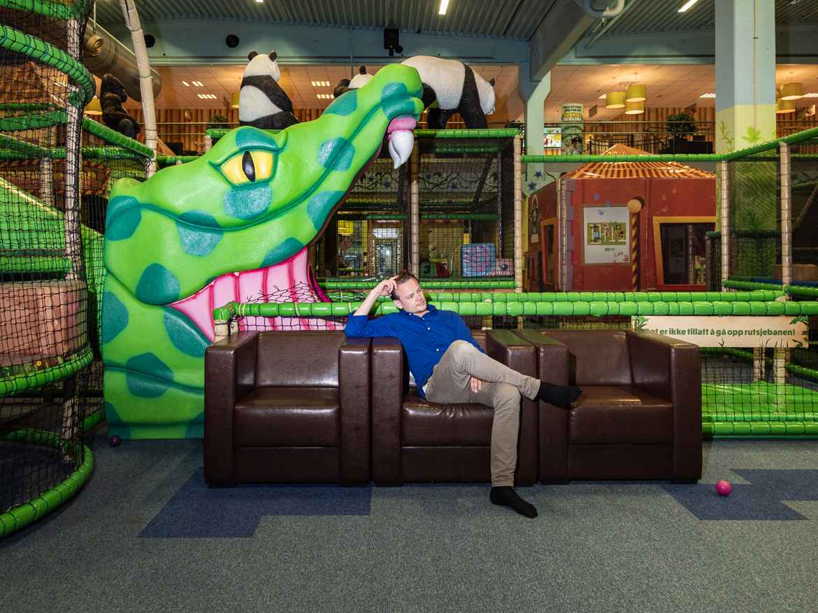 Man sitting in brown leather sofa attending children at an indoor playground. Behind him is an enormous crocodile head with its mouth gaping over him.