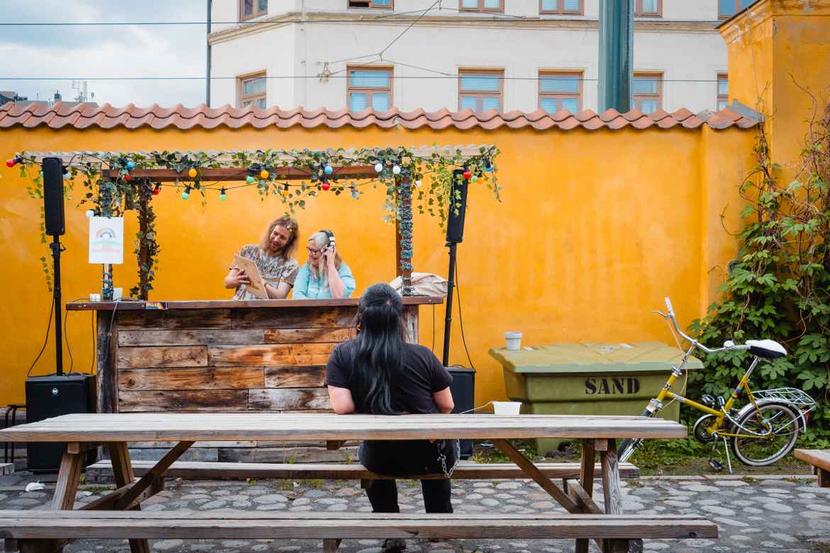 Two prog rock DJs in wooden DJ booth preoccupied with exploring each other's recommendations while a sole audience member leans back on a bench observing them.