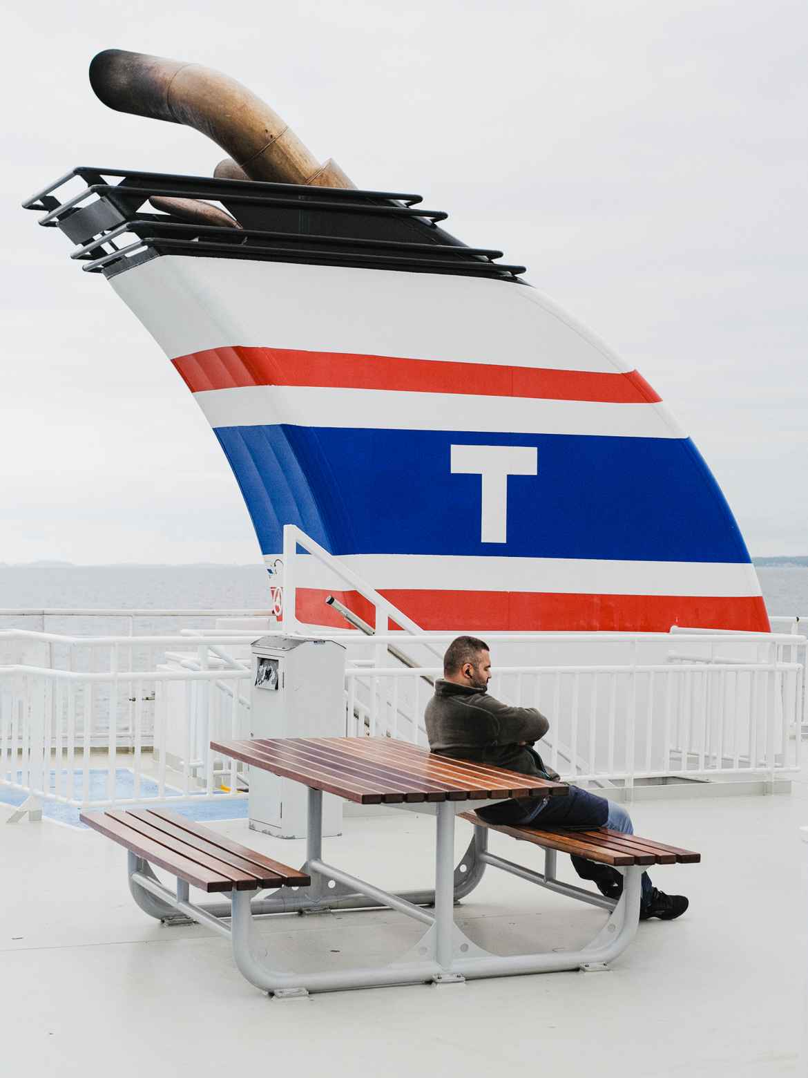 Man leaning back on a bench on a ferry with a large chimney in the background shaped opposite to his posture.