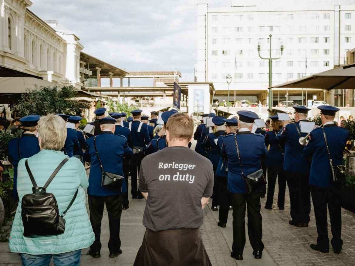 Crowd with marching band photographed from behind. Foreground shows a man's back with a T-shirt that says "Plumber on duty" in Norwegian.