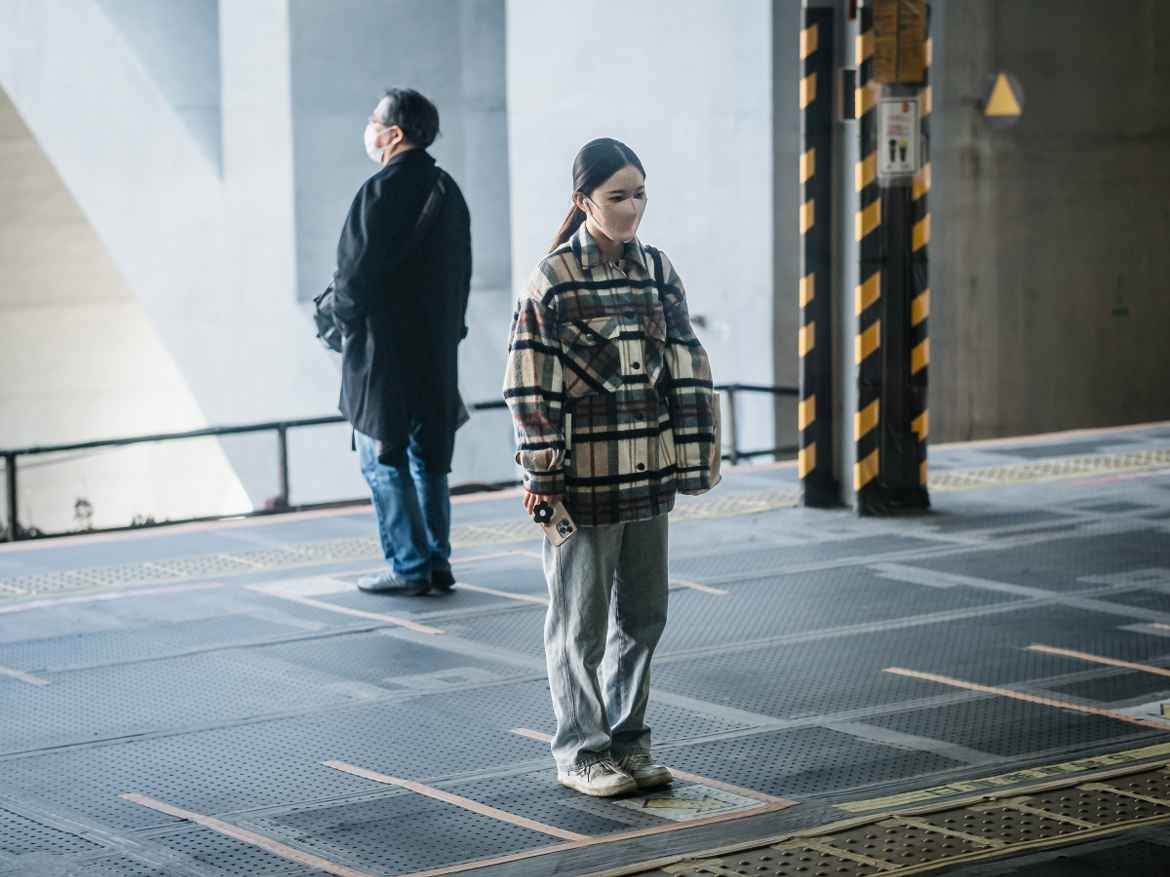 Two Japanese metro passengers, backs to each other, waiting for the train, dilligently standing on the correct markings on the platform.